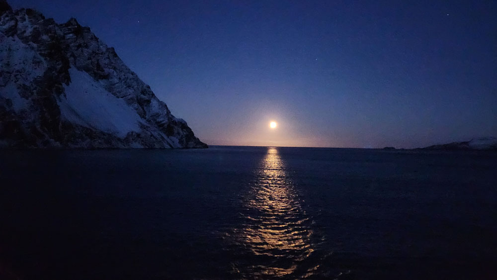 A full moon setting over the ocean with snow on it.